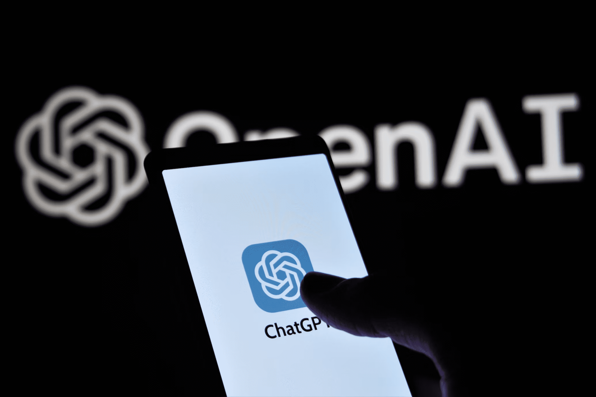 Foto com uma mão segurando um celular com o aplicativo do ChatGPT em destaque na tela. De fundo, num background preto, tem a logo do ChatGPT mais o texto OpenAI.
