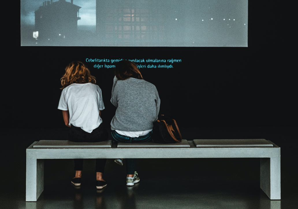 Foto mostrando duas pessoas vendo um material audiovisual com legendas numa sala.