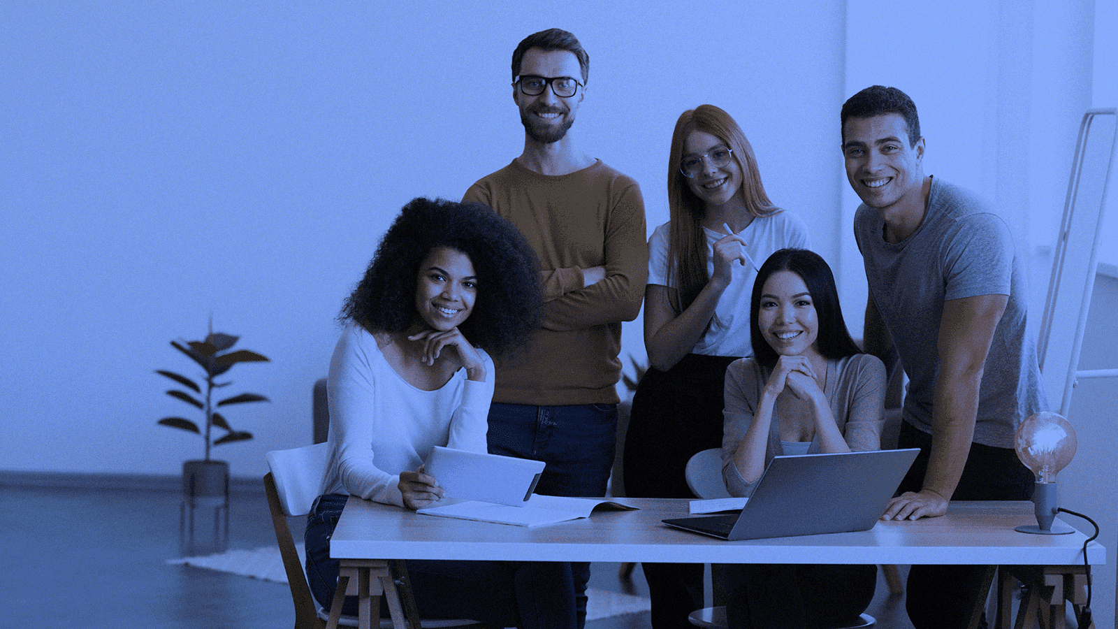 Co-Marketing - Pessoas reunidas numa mesa, pousando e sorrindo para a foto tirada. De fundo, há uma parede branca e uma planta no jarro.