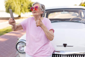 idosa encostada num carro, com uma blusa e oculos rosa, com o celular na mão, tirando uma selfie.