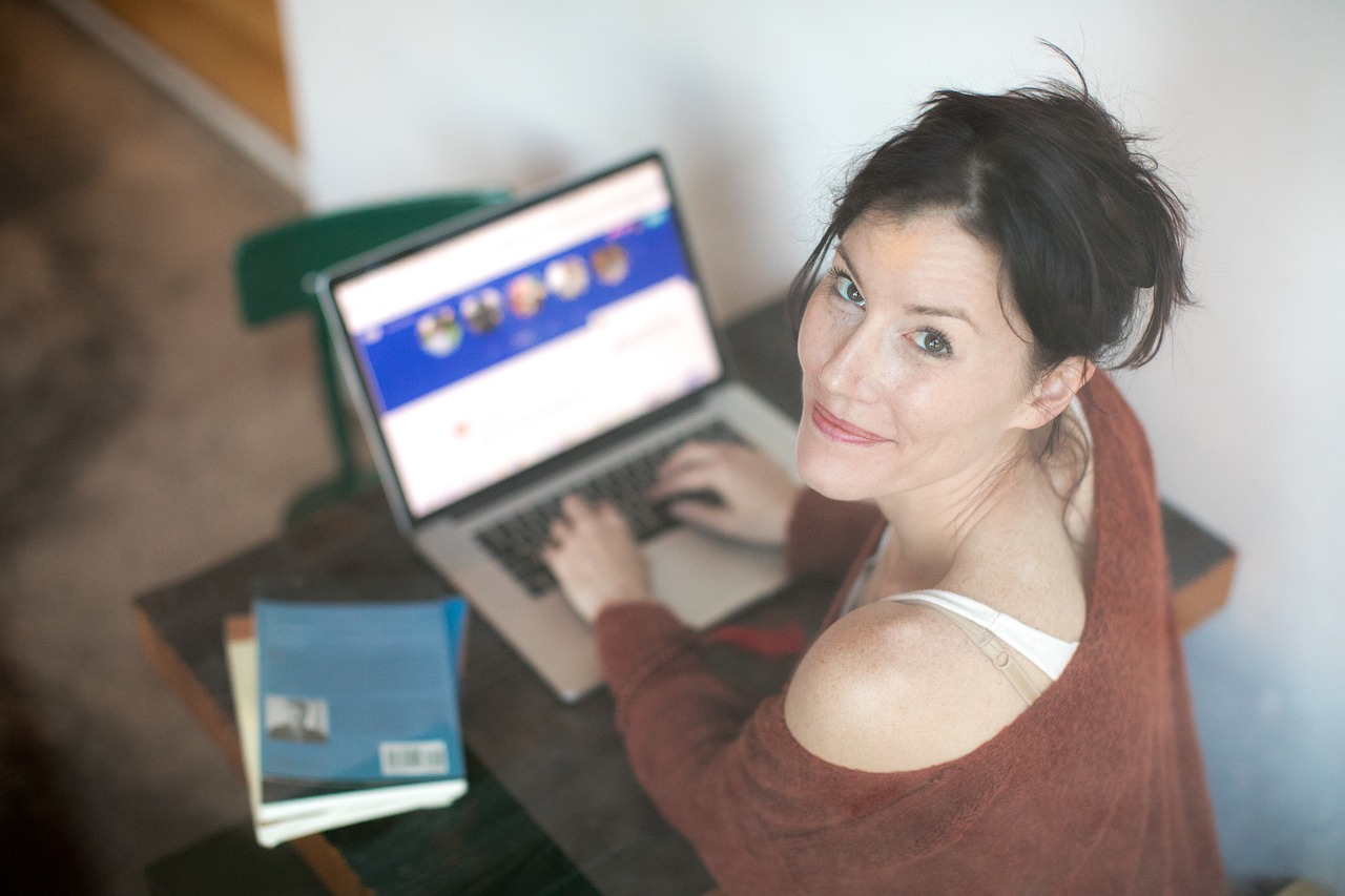 interação com seguidores - Imagem de uma mulher branca, de cabelo castanho e preso, sentada num ambiente com o computador no colo, posicionada para a foto olhando para cima. Ela usa uma camisa de lã marrom com uma branca por baixo.