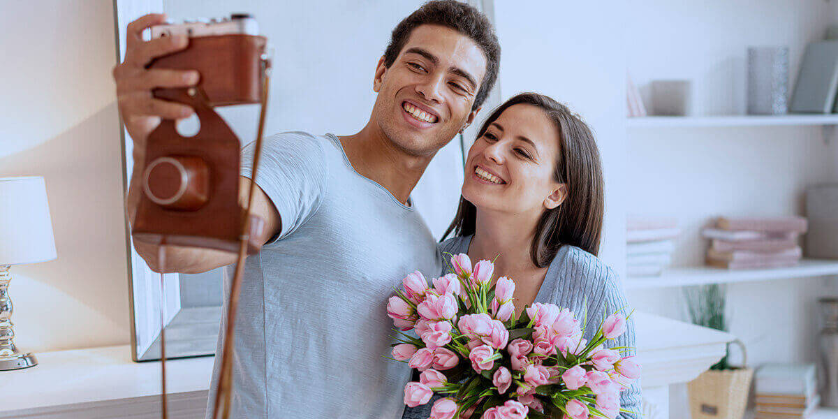 Dia dos namorados - Imagem ilustrando o tema com um casal, numa sala de estar de fundo, tirando uma foto numa câmera modelo antigo. O homem é pardo, de cabelo castanho, segurando a câmera, sorrindo e abraçando uma mulher. Ela é parta, de cabelo castanho, está usando uma camisa cinza e segurando um buquê de flores.