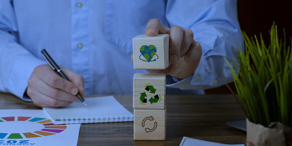 Greenwashing - Imagem ilustrativa do tema do artigo onde tem mesa com os braços de um homem apoiados onde uma mão segura uma caneta sobre um bloco de notas e, a outra mão, está segurando um dado com o símbolo de um coração feito com a ilustração do planeta terra e, esse cubo, está em cima de mais dois onde tem o símbolo da reciclagem e reutilização.