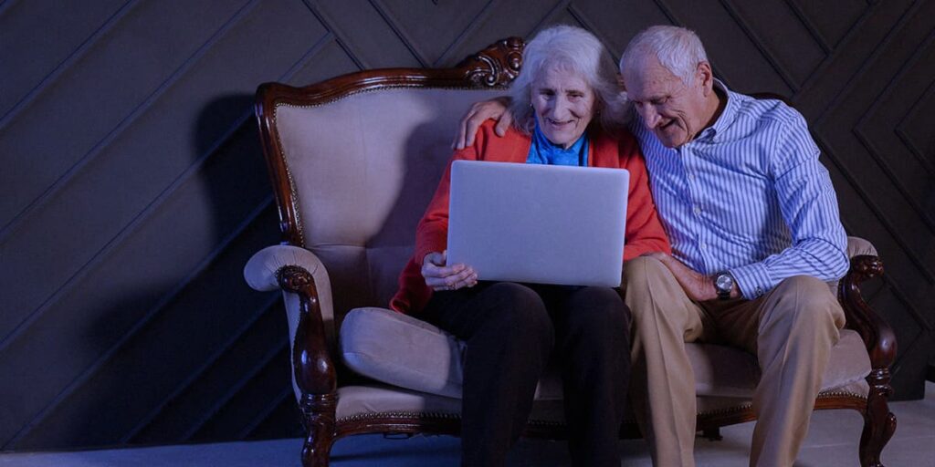 Um casal de idosos sentados no sofa, com o notebook no colo, olhando para a tela.