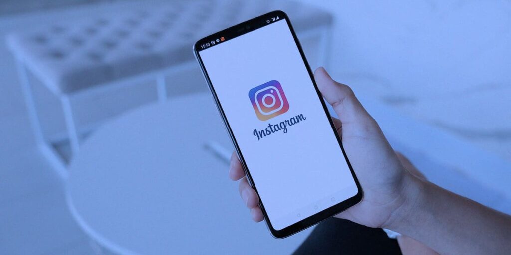 campanhas no Instagram - na imagem tem uma mão, segurando um celular com a logo e nome do Instagram abertos na tela. De fundo, tem uma sala de estar, onde aparece um sofá pequeno e uma mesa de centro.