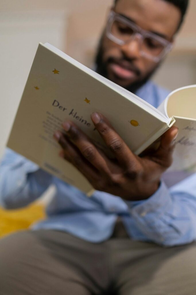 a imagem que ilustra o tópico tem um homem negro, usando uma camisa de botão azul com uma calça social bege, óculo de armação clara, segundo e lendo um livro. De fundo, o ambiente, é um quarto. 