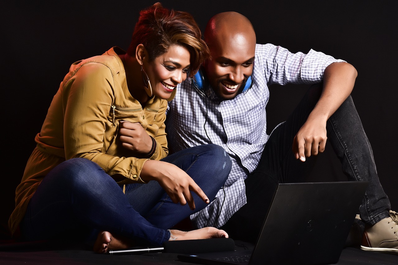 a imagem que ilustra o tópico, tem um casal sentado num cômodo de fundo escuro, aparentemente, no chão, olhando para um computador. A mulher é negra, usa calça jeans e uma camisa de botão amarelo escuro. O homem, é negro, careca, usa uma camisa de botão azul e uma calça escura.