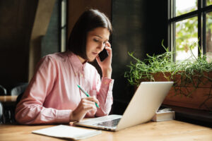 Mulher com blusa rosa, sentada numa mesa, falando ao celular, e com o notebook aberto, enquanto resolve problemas.