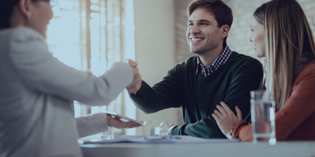 Relacionamento com o Cliente - a imagem que ilustra o tema tem 3 pessoas: duas mulheres e um homem. Uma das mulheres está usando um blazer branco, olhando para o homem e apertando a mão dele. O homem é branco de cabelo claro, está usando uma camisa preta e sorrindo para a primeira mulher. A segunda mulher, está ao lado do homem, usando uma blusa marrom, segurando o braço dele e ela é branca de cabelo liso claro.