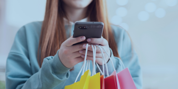 Consumidor 5.0 - a imagem que ilustra o tema é de uma mulher branca, de cabelos ruivos, usando uma camisa de manga azul, segurando o celular e sacolas de compras.