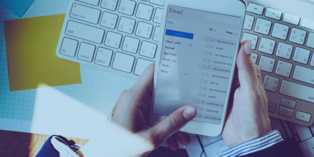 Abertura de email - a imagem que ilustra o tema tem um teclado de computador, de fundo, sobre uma mesa branca onde também aparece um post it amarelo e, uma mão de uma pessoa, segurando um celular onde tem uma caixa de email sendo aberta.