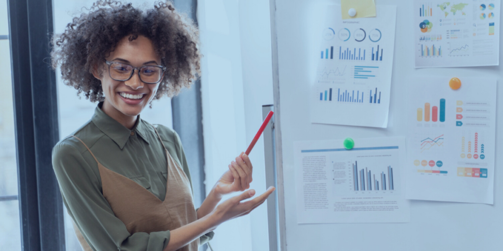 Estrategista de Marketing - a imagem que ilustra o tema tem uma mulher negra, de cabelo cacheado, usando óculos, com uma camisa verde escuro de botão e, por cima, uma blusa regata verde claro. Ela está numa sala de reunião, apontando para um quadro e apresentando gráficos que estão exibidos num quadro branco.