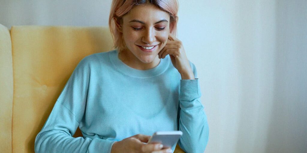 responder mensagens diretas - a imagem que ilustra o tema tem uma mulher, branca, de cabelo rosa, usando uma camisa de manga longa azul, sentada numa cadeira amarela, segurando o celular e sorrindo.