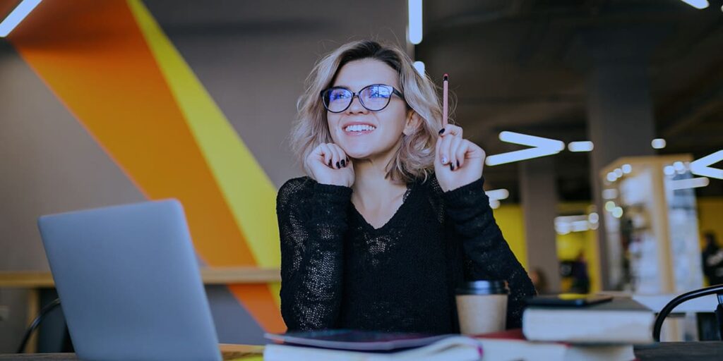 Marketing sem rosto - a imagem que ilustra o tema tem uma mulher branca, de cabelo loiro, usando óculos preto, blusa de manga longa vazada preta, sentada segurando um lápis para cima e sorrindo, dando o sentimento de que teve uma ideia. De fundo, tem um ambiente corporativo, aparentemente, um coworking.