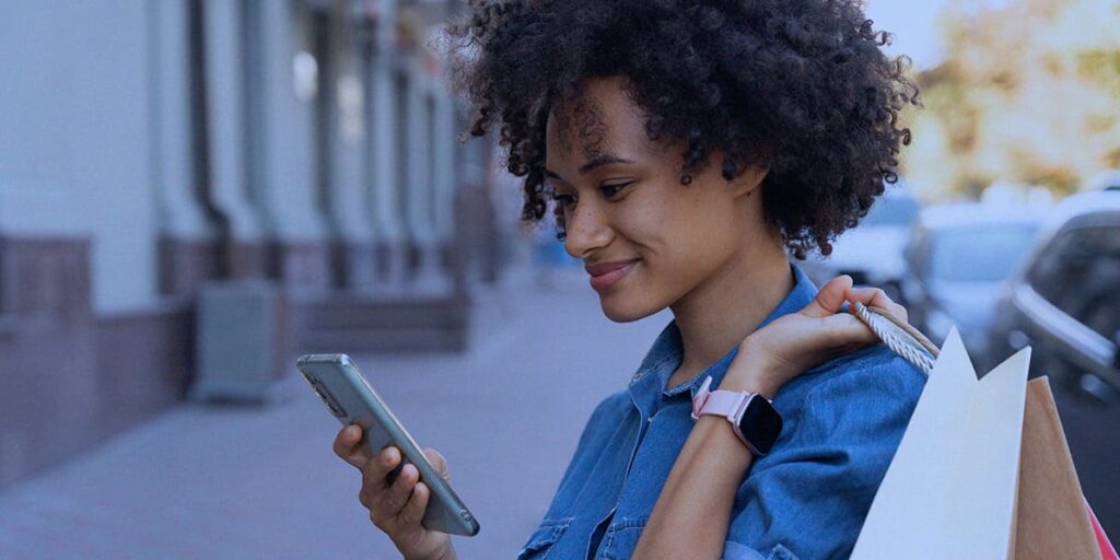 comportamento do consumidor - a imagem que ilustra o tema tem o fundo de uma rua e, em destaque, tem uma mulher negra, de cabelo curto cacheado, usando uma camisa azul, segurando sacolas de compras, segurando e olhando para um celular.