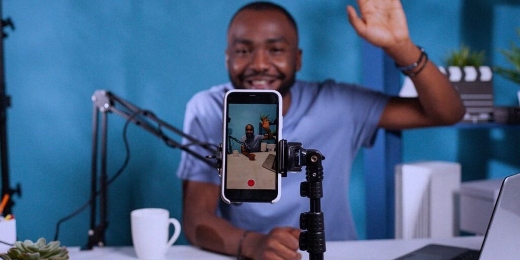 Lives para amigos próximos - a imagem tem um cenário de fundo azul, com plantas e uma claquete de gravação. No meio, tem um homem negro usando uma camisa azul, de cabelo curtinho, acenando para um celular que está num tripé com a tela de gravação aberta.