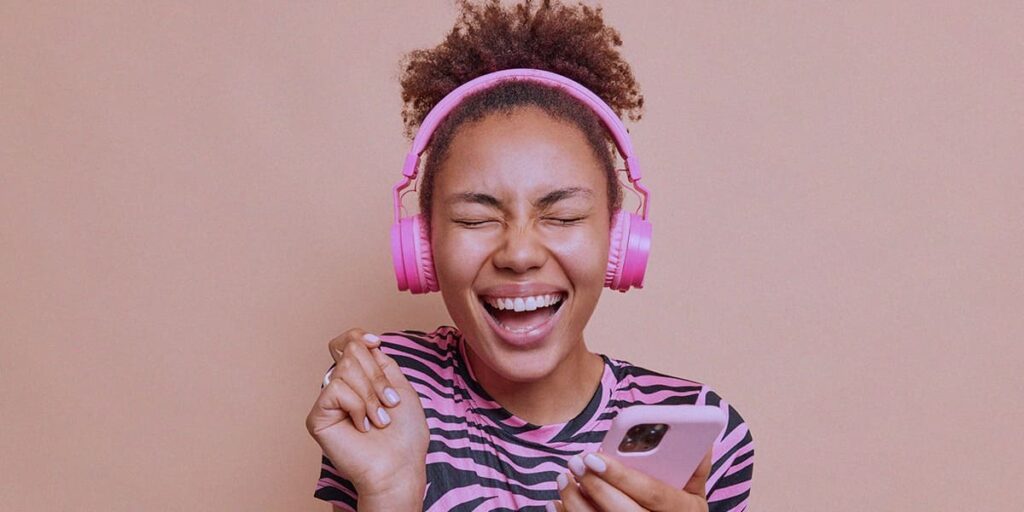 Melhores músicas para Reels - A imagem que ilustra o tema tem o fundo rosa, no meio, em destaque, tem uma mulher negra, de cabelo cacheado preso, usando uma camisa de manga curta com listras pretas e brancas, segurando um celular, usando um headphone e sorrindo fazendo expressão de feliz enquanto ouve música.