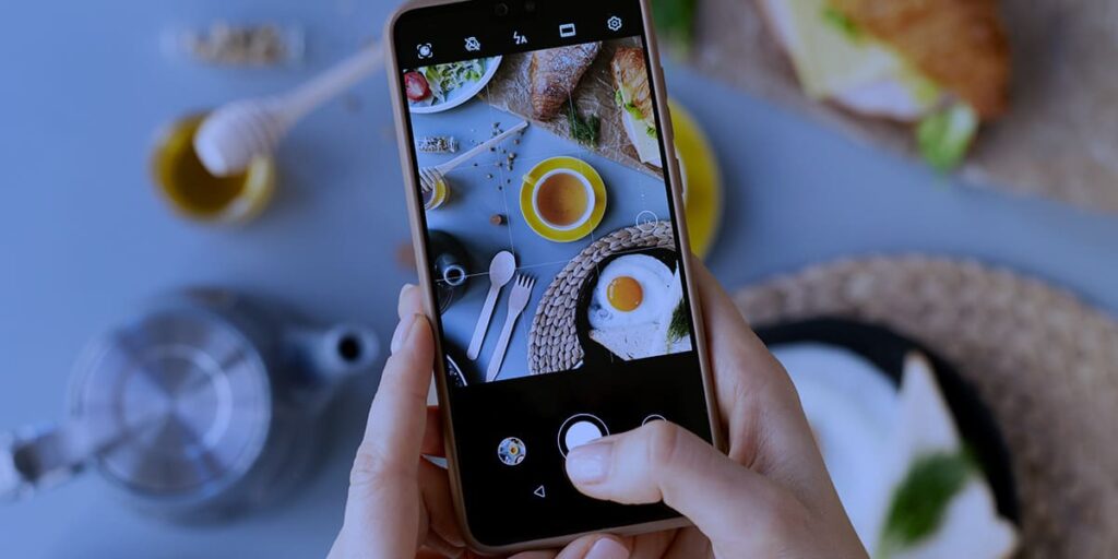 variação de conteúdo no Instagram - a foto que ilustra o ema tem o fundo de uma mesa branca com um café da manhã e, em destaque, tem duas mãos de uma pessoa branca, segurando um celular e fazendo um enquadramento da mesa para captação de uma foto.