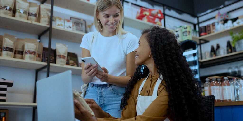 Jornada de compra do Consumidor 5.0 - a imagem que ilustra o tema tem duas mulheres: uma branca de cabelo liso, loiro, usando camisa branca e calça jeans segurando um celular. E, a outra mulher, é negra, usando uma camisa num tom terroso, de cabelo longo cacheado, usando um avental branco sentada em frente a um computador também segurando um celular e olhando para a outra mulher sorrindo. De fundo, tem uma loja que vende alimentos e bebidas.