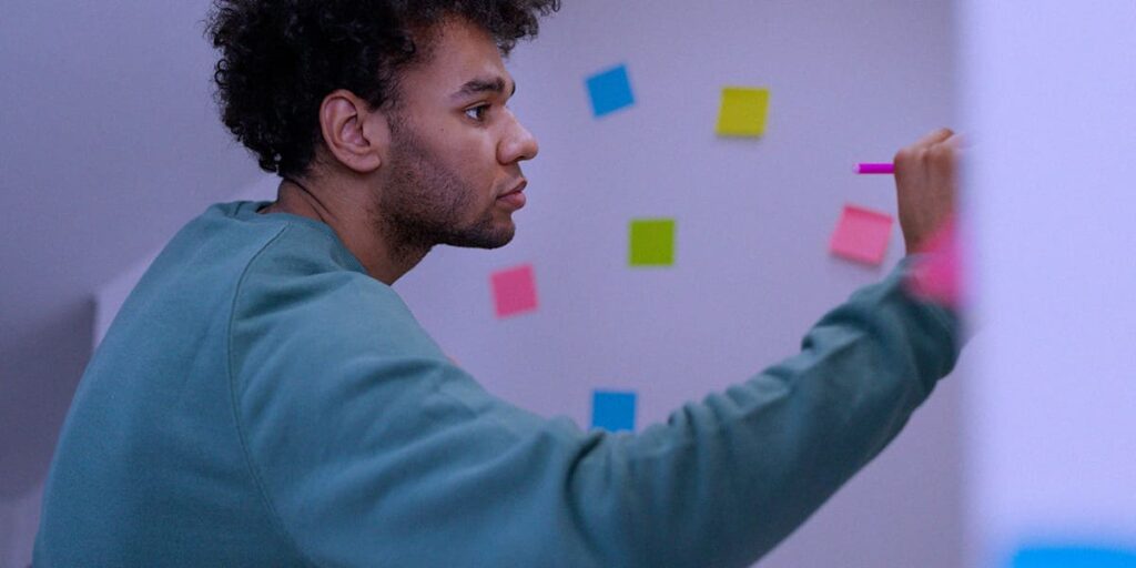 Metodologia Sprint - A imagem que ilustra o tema tem um homem, negro, de cabelo curto cacheado, usando uma camisa de manga longa escrevendo num quadro onde tem diversos post its colados