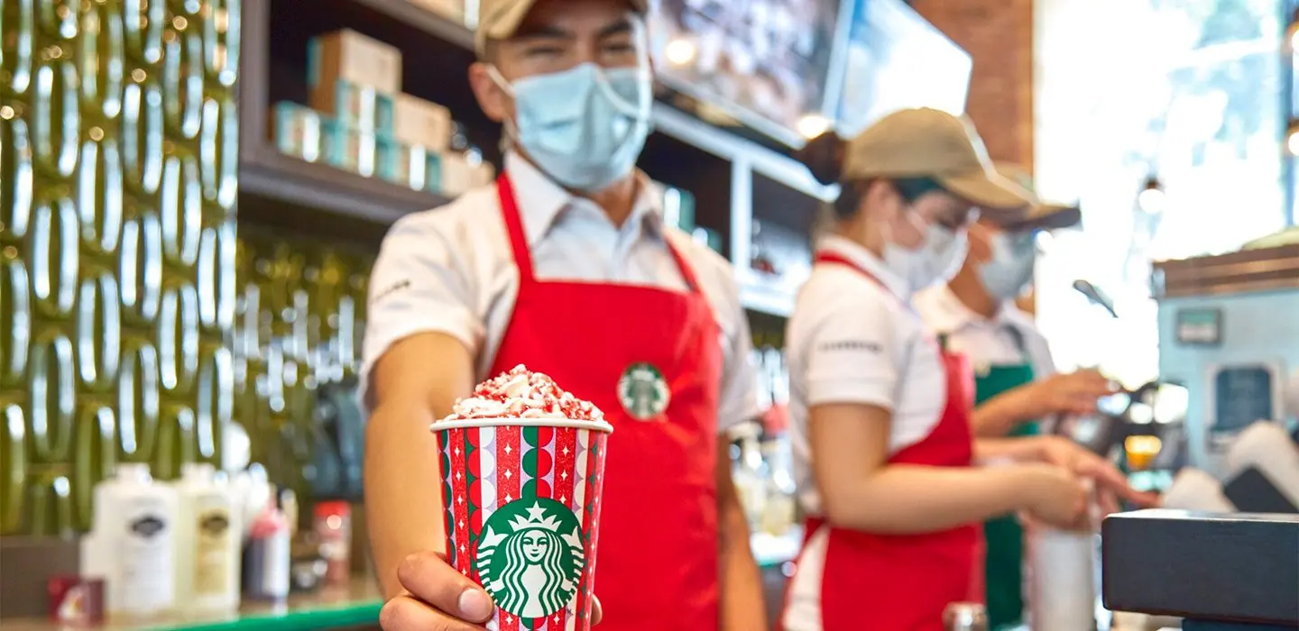 A imagem tem dois atendentes da Starbucks mostrando itens personalizados da marca para o Natal.