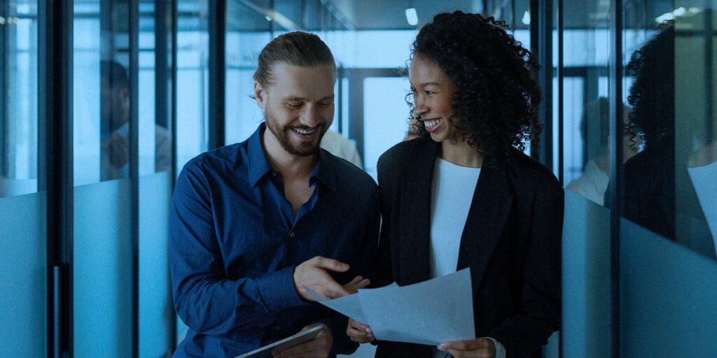 Diagnóstico na Consultoria Gratuita - A imagem que ilustra o tema tem o corredor de um escritório, onde estão duas pessoas, um homem branco de cabelo liso, loiro, usando uma camisa de botão azul escuro, sorrindo e apontando para o papel que a mulher, ao seu lado, está segurando. Ela é negra, tem cabelo cacheado curto, está usando um blazer preto e uma camisa branca por baixo.