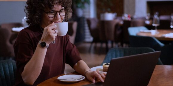 Proposta de consultoria gratuita - A imagem que ilustra o tema temo fundo de uma cafeteria e, em destaque, uma mulher parda, usando uma camisa marrom, sentada em frente o computador e bebendo café o chá, o líquido que estiver dentro do recipiente.