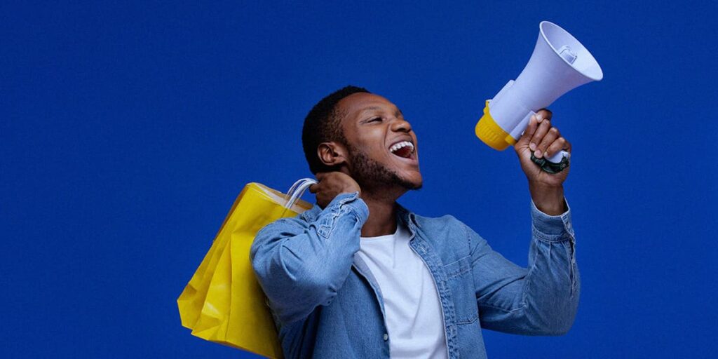 Pacotes Promocionais - A imagem que ilustra o tema tem o fundo azul escuro com um homem, negro, posicionado de forma centralizado, segurando um megafone e uma sacola amarela de compra. Ele está vestindo uma camisa branca e, por cima, uma camisa de botão azul.