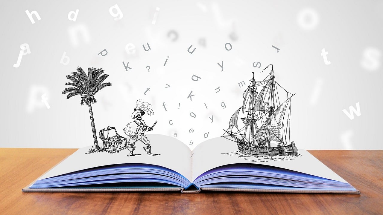 A imagem que ilustra o parágrafo tem o fundo branco com uma mesa de madeira onde aparece um livro pop up em cima dela aberto e, nas páginas, aparecem várias letras voando um coqueiro, uma ilha com tesouro, um pirata e um navio.