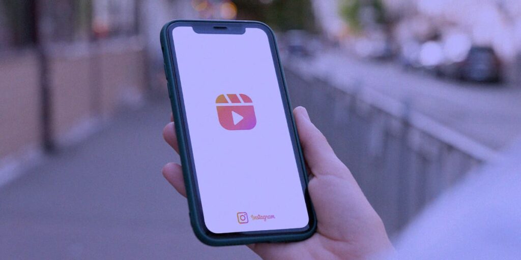 Carrossel que aparece no feed do reels - a imagem que ilustra o tema tem de fundo uma rua e, em destaque, aparece uma mão de uma pessoa branca, segurando um celular com o ícone do reel aberto na tela na cor rosa.
