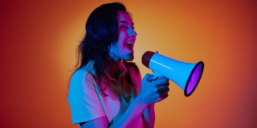 Marketing de Retenção - A imagem que ilustra o tema tem o fundo laranja com uma vinheta preta nas bordas e, no meio, tem uma mulher, de cabelo solto longo, usando uma camisa de manga média branca, segurando um mega fone e a boca aberta, como se estivesse falando através dele.