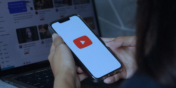 Mulher com o celular na não, abrindo o youtube e com o notebook aberto na mesa e o Short do youtube aberto na tela