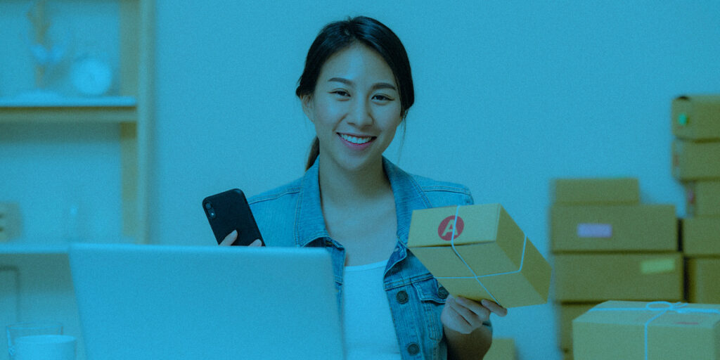 Mulher com o notebook aberto na mesa, varias entregas embaladas para cross-sell e o celular na mão.